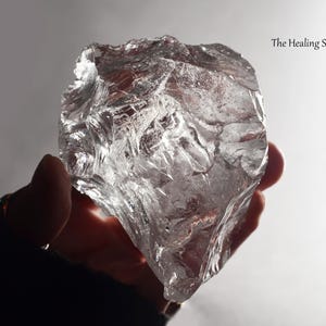 May include: A large, clear, irregularly shaped crystal is held in a hand. The crystal has a rough, textured surface and appears to be translucent. The text "The Healing Sea Wolf" is in the upper right corner.