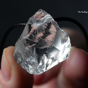 Op de afbeelding: Een heldere, ruw geslepen kristallen hanger die tussen vingers wordt gehouden. Het kristal is facetgeslepen, reflecteert licht en lijkt doorschijnend. Een zwart koord is aan het kristal bevestigd. The Healing Sea Wolf staat rechtsboven.