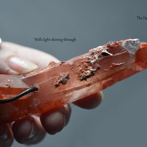 Op de afbeelding: Een rood-oranje kristal met een ruw, getextureerd oppervlak wordt vastgehouden. Het kristal heeft een zwart koord. De tekst "With light shining through" en "The Healing Sea Wolf" zijn zichtbaar.