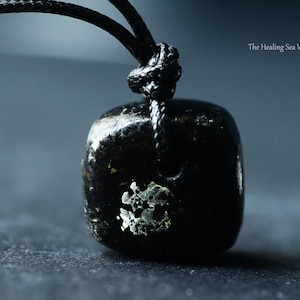Puede incluir: Un colgante de piedra oscura, pulida y con forma de cubo, suspendido de un cordón negro. La piedra presenta una inclusión metálica de color plateado irregular en su parte frontal. El texto "The Healing Sea Wolf" aparece en la esquina superior derecha.