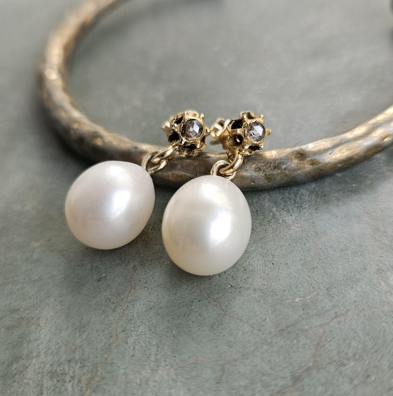 May include: A pair of gold-toned earrings with a pearl drop. The earrings have a small, textured top with a clear stone. The pearls are oval-shaped and have a lustrous, white finish. The earrings are displayed on a gold-toned, textured surface.
