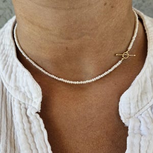 May include: A delicate necklace featuring a single strand of small, white, round seed pearls. The necklace is secured with a gold-toned metal toggle clasp, consisting of a circular ring and a horizontal bar, positioned off-centre on the right side.