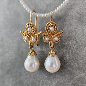 Puede incluir: Un par de pendientes colgantes dorados con una secci&oacute;n superior texturizada y ornamentada, acentuada con tres peque&ntilde;as perlas blancas. Cada pendiente suspende una perla blanca lisa en forma de l&aacute;grima desde un casquillo dorado decorativo.