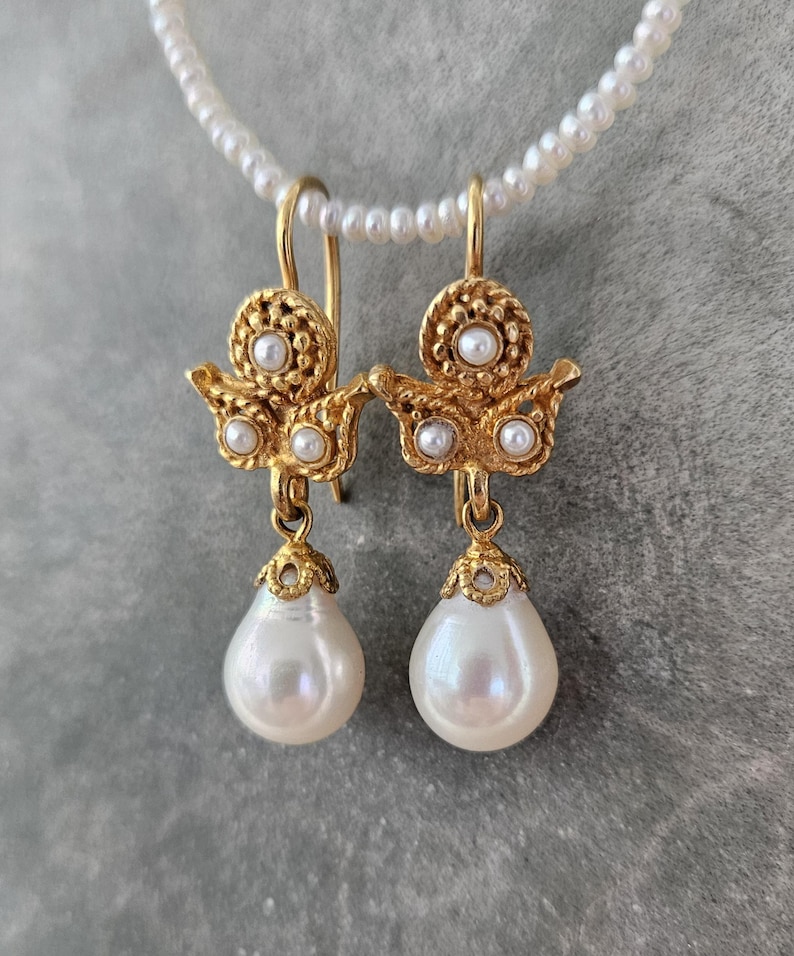 Puede incluir: Un par de pendientes colgantes dorados con una secci&oacute;n superior texturizada y ornamentada, acentuada con tres peque&ntilde;as perlas blancas. Cada pendiente suspende una perla blanca lisa en forma de l&aacute;grima desde un casquillo dorado decorativo.