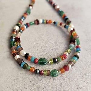 Op de afbeelding: Een meerstrengs ketting met een verscheidenheid aan kleine, gefacetteerde, veelkleurige edelsteenkralen afgewisseld met witte parels. De kralen bevatten tinten groen, rood, blauw, oranje en zwart, geaccentueerd door kleine goudkleurige metalen tussenstukjes.
