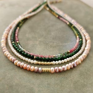 Może przedstawiać: Cztery warstwowe naszyjniki z różnymi rodzajami koralików. Górna warstwa to mieszanka małych, jasnych koralików. Druga warstwa to zielone koraliki z różowymi i złotymi akcentami. Trzecia warstwa to białe perły. Dolna warstwa to różowe perły ze złotymi koralikami.