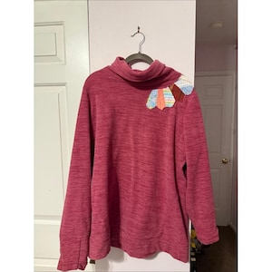May include: A long-sleeved, heathered magenta turtleneck sweater featuring a decorative, fan-shaped appliqué on the left shoulder. The appliqué is composed of multiple fabric segments in various pastel colors and patterns, including plaid and gingham.