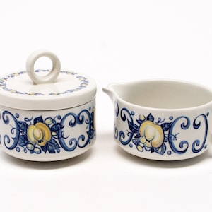 May include: A white ceramic creamer and sugar bowl set with a blue and yellow floral design. The creamer has a handle and the sugar bowl has a lid with a ring handle.