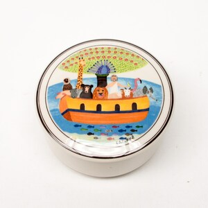 May include: A round ceramic trinket box featuring a colourful illustration of a boat filled with animals, a peacock, and a tree. The boat is orange and yellow, and the animals are a variety of colours. The background is blue with white waves.