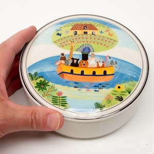 May include: A round ceramic trinket box with a colourful illustration of animals on a boat sailing on a blue sea. The lid features a green island with a house and a peacock.