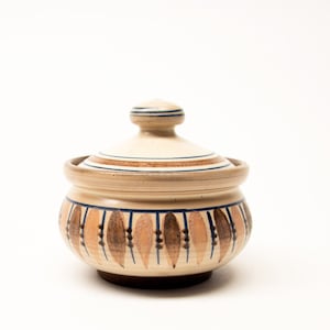 May include: A round ceramic lidded jar with a cream-coloured base. The body features a repeating pattern of vertical brown leaf-shaped motifs separated by thin blue lines. The lid has a matching knob handle with blue and brown circular bands.