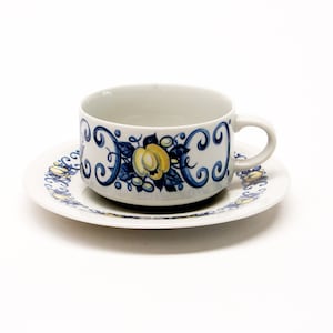 May include: A white ceramic teacup and saucer set. The cup and saucer are decorated with a blue and yellow floral design and a scroll pattern. The cup has a handle and sits on the matching saucer.