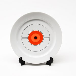 May include: A white ceramic plate with a black and orange concentric circle design. The plate is on a black stand.