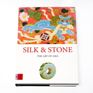 May include: A hardback book titled SILK & STONE THE ART OF ASIA. The cover features a vibrant orange textile print with a blue mythical creature and white flowers, and a central image of a circular, pale green carved jade-like stone object.