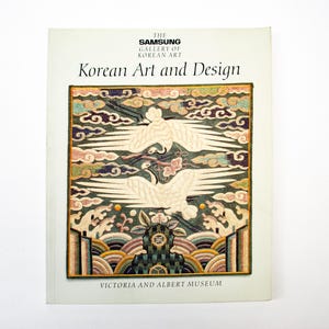 以下が含まれることがあります： 「The Samsung Gallery of Korean Art: Korean Art and Design, Victoria and Albert Museum」というタイトルのソフトカバー本。表紙には、様式化された波とカラフルな雲の上を飛ぶ2羽の白い鶴の刺繍テキスタイルイラストが描かれています。