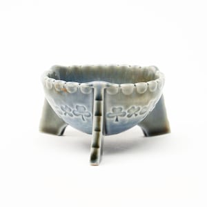 May include: A small, three-legged ceramic bowl featuring a glossy, variegated blue and grey glaze. The exterior is decorated with a repeating embossed shamrock pattern around the centre, and the rim has a scalloped edge design.