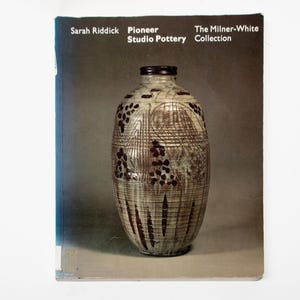 May include: A book titled Sarah Riddick Pioneer Studio Pottery The Milner-White Collection featuring a tall, cylindrical ceramic vase with a textured, light-coloured surface, dark brown decorative markings, and a dark rim on a dark grey background.