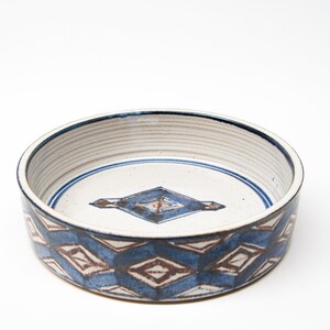 May include: A white ceramic bowl with a blue and brown geometric pattern. The bowl has a wide rim and a slightly rounded bottom.