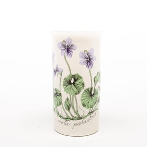 May include: A white ceramic vase with a botanical illustration of purple violets, green leaves, and the text "viola palustris." The vase has a cylindrical shape and is set against a white background.