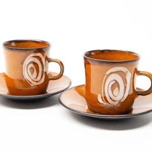 May include: Two brown ceramic coffee cups and saucers. Each cup has a white spiral design and a handle. The set is on a white background, highlighting the warm tones of the pottery.