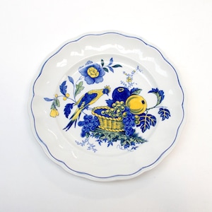 May include: White ceramic plate with a scalloped edge and a blue border. The plate features a colourful illustration of a bird, fruit, and flowers in shades of blue, yellow, and green. A decorative piece for serving or display.