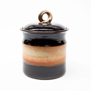 May include: A ceramic storage jar with a lid. The jar is dark brown at the top and base, with a tan and reddish-brown band in the middle. The lid has a loop handle. The jar is likely used for storage.