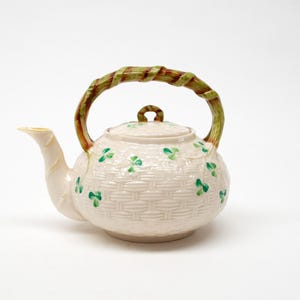 May include: A cream-coloured ceramic teapot featuring a raised basket-weave texture across the body. The teapot is decorated with small, hand-painted green shamrock motifs and includes a handle shaped like a textured, brown and green bamboo stalk.