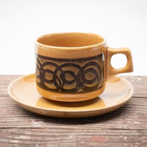 May include: A tan ceramic cup and saucer set featuring a dark brown band with a repeating circular loop pattern around the centre of the cup. The cup has a square-shaped handle and a smooth, glazed finish, resting on a matching circular saucer.