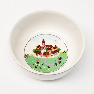 May include: A white ceramic bowl featuring a circular illustration at the bottom. The scene depicts a small village with houses, a church, and a green field with chickens and two figures. The word LAPLAU is printed in the lower right corner.