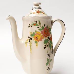 May include: A white ceramic coffee pot with a floral design. The pot has a black trim and a lid with a knob. The flowers are pink, yellow, and orange.