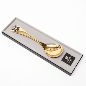 May include: A gold-toned spoon with a dark blue butterfly motif on the handle, presented in a rectangular black box with a grey interior lining. A small black square label with a gold crown logo and text is positioned next to the spoon bowl.