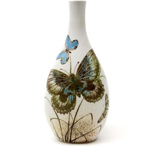 May include: A white ceramic vase with a narrow neck and a bulbous body. It features detailed butterfly illustrations in blue, green, and brown, with fine plant stems. The design is elegant and inspired by nature. The vase is approximately 20 cm tall.