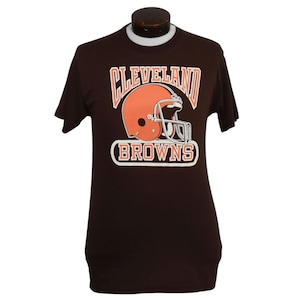 May include: A dark brown short-sleeve t-shirt featuring a graphic of an orange football helmet. Above the helmet, the word CLEVELAND is printed in white with an orange outline. Below the helmet, the word BROWNS is printed in white inside an oval.