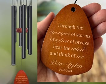 May include: A wind chime with black tubes and a wooden top and accents. A wooden teardrop-shaped pendant with the text "Through the strongest of storms or softest of breeze hear the wind and think of me Peter Dylan 1940-2024."