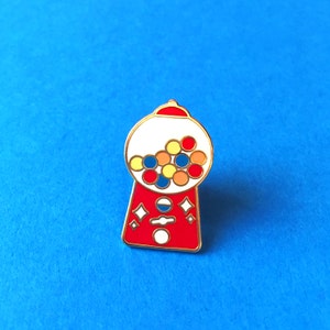 May include: A red and white gumball machine enamel pin with gold trim. The pin features a clear bubble filled with colorful gumballs. The pin has a classic design with diamond and circle accents.