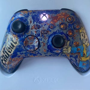 May include: Custom Xbox controller with a Fallout-themed design. The controller features a blue, orange, and gray color scheme with the word "Fallout" printed on the left side. The Xbox logo is illuminated.