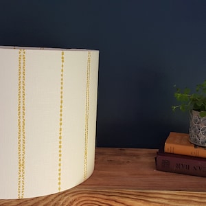 May include: A white lampshade with yellow vertical stripes. The lampshade is on a wooden shelf with a small potted plant and a stack of books.
