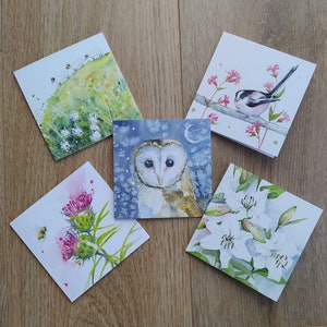 May include: Five square watercolour art prints featuring nature scenes. The prints depict an owl, a bird on a branch, sheep on a hill, a bee and thistle, and white lilies. The art is displayed on a wooden surface.