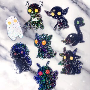 May include: A collection of glittery stickers featuring various fantasy creatures. The stickers include a ghost, a skeletal figure, a goblin, a dragon, a Loch Ness Monster, a furry creature, a moth, and a horned demon. Each sticker has a unique design and color scheme.