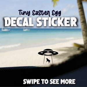 May include: A black decal sticker with the text "Tiny Easter Egg DECAL STICKER" in white and blue. The decal features a UFO with a dog silhouette, set against a beach scene. The text "SWIPE TO SEE MORE" is at the bottom.