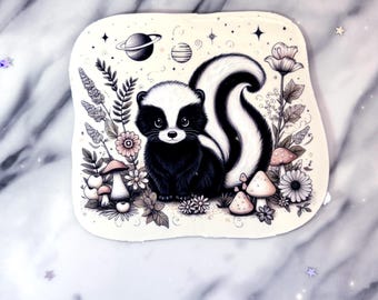 May include: A whimsical illustration of a skunk surrounded by flowers, mushrooms, and celestial elements. The skunk is depicted in black and white with a fluffy tail. The artwork has a soft, pastel colour palette and a dreamy aesthetic.