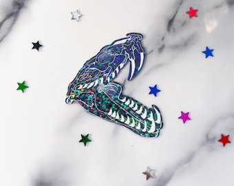 Può includere: Un adesivo olografico di un teschio di serpente su sfondo bianco. L'adesivo è circondato da stelle colorate.