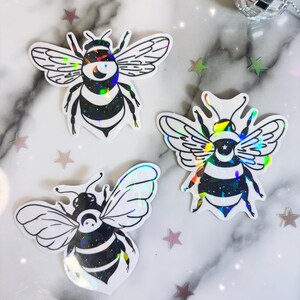 May include: Three iridescent holographic bee stickers with black and white outlines. The bees have a crescent moon and star design on their bodies.