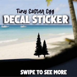 May include: Black decal sticker with two pine trees. The text "Tiny Easter Egg DECAL STICKER" is above the trees. The background shows a beach scene with boats and palm trees. The text "SWIPE TO SEE MORE" is at the bottom.