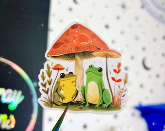 Könnte beinhalten: Ein Aufkleber mit zwei Fröschen, die unter einem roten und weißen Pilz sitzen. Der Frosch links ist gelb und der Frosch rechts ist grün. Der Pilz hat weiße Punkte und einen braunen Stiel.