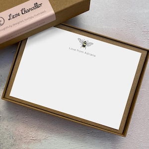 May include: A set of white stationery cards inside a brown cardboard box. Each card features a small, detailed illustration of a bee in black and yellow, with the text "Love from Adriana" printed in a simple black font below the bee graphic.
