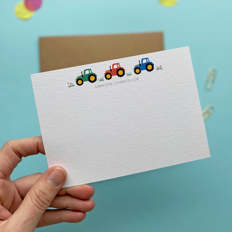 May include: A white card with a line of four toy tractors in green, red, and blue. The tractors are driving on a green field with small flowers. The text "GRAYSON CHANCELLOR" is printed below the tractors.