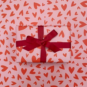 Pu&ograve; includere: Una confezione regalo avvolta con un nastro e un fiocco bordeaux &egrave; appoggiata su carta da regalo a forma di cuori rossi su sfondo rosa. La confezione regalo &egrave; avvolta in carta con un motivo a cuore simile.