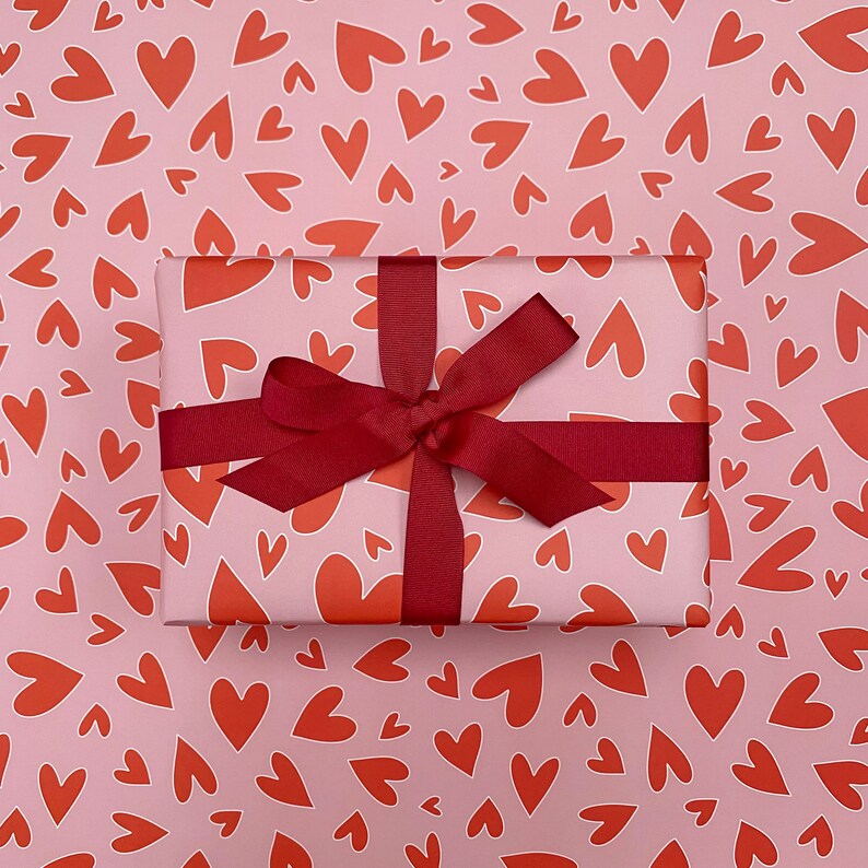 Pu&ograve; includere: Una confezione regalo avvolta con un nastro e un fiocco bordeaux &egrave; appoggiata su carta da regalo a forma di cuori rossi su sfondo rosa. La confezione regalo &egrave; avvolta in carta con un motivo a cuore simile.