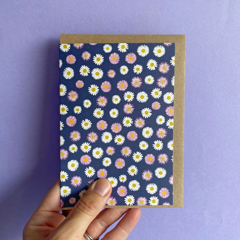 Pode incluir: Um cart&atilde;o de felicita&ccedil;&otilde;es em branco com um fundo azul marinho e um padr&atilde;o de margaridas brancas e rosas. O cart&atilde;o tem um envelope castanho.